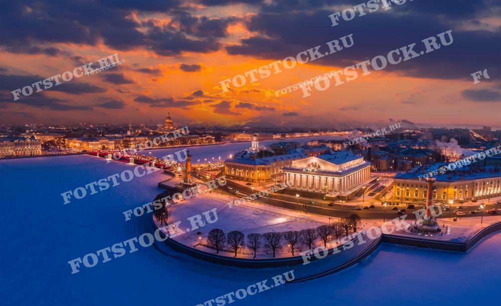На этой панорамной фотографии запечатлен вечерний Санкт-Петербург в зимнее время. Город покрыт снегом, что добавляет ему очарования и тишины. Освещение зданий и улиц создаёт тёплый свет в контрасте с прохладными синими оттенками снега и неба. Вдалеке видны силуэты известных достопримечательностей. Это захватывающий вид открывает красоту городского пейзажа во время заката, подчёркивая его историческое наследие и современное очарование.